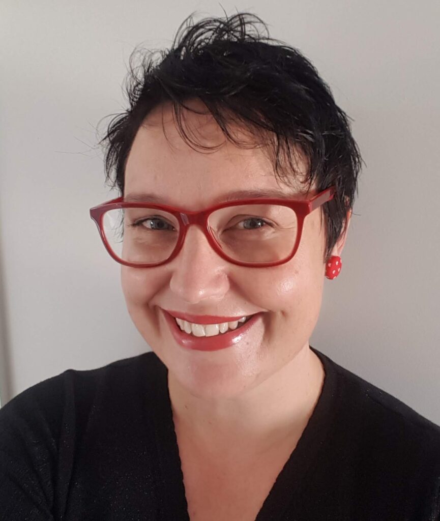 Woman smiling. Red glasses and red earrings. She has black hair and blue eyes. The image introduces the writer - Lisa.